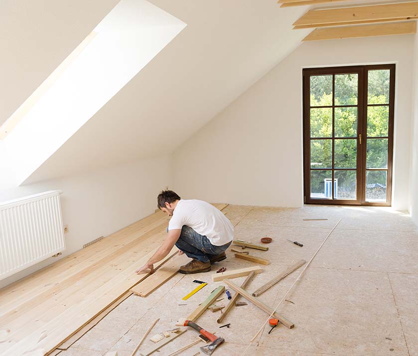 A man installing wooden floorboards in an attic, surrounded by tools and materials.